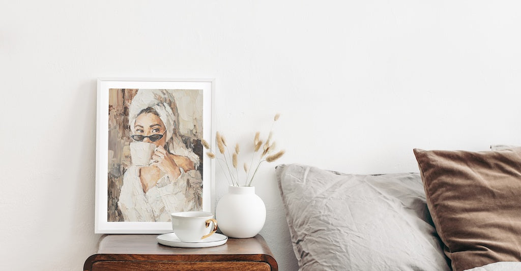 Framed print of a portrait of a woman drinking a coffee pictured in a neutral bedroom. 