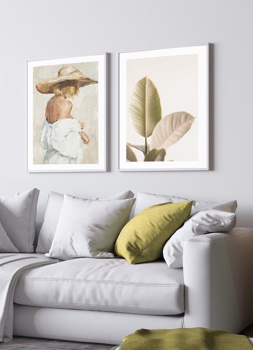 beautiful gallery wall with a painted poster print of a female wearing a sunhat and a botanical green print pictured in a grey and green living room. 